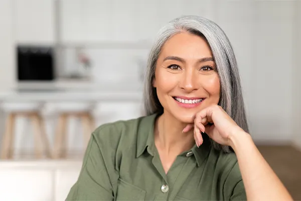 Menopause-Clinic An asian woman with long gray hair and a green shirt sitting and smiling on her couch, happy with her menopause care from [NAME} in Boca Raton.