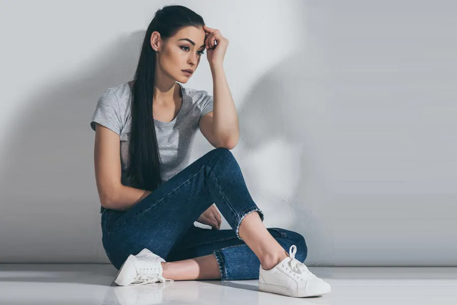 PCOS-Doctor A young brunette woman in a gray t-shirt and jeans sits against a gray wall with her head resting on her hand and a distressed expression. Get help for Polycystic Ovarian Syndrome (PCOS) from Richard Conlen, MD, FACOG in Boca Raton.