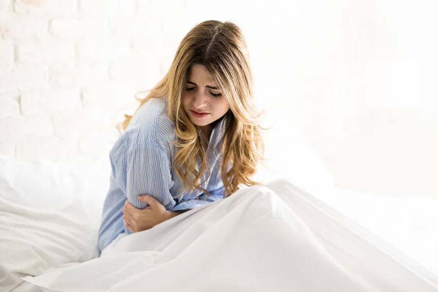 PMS-Therapy A woman with long blonde hair sits in her bed holding her stomach in pain, considering getting care for PMS from Richard Conlen, MD, FACOG in Boca Raton.